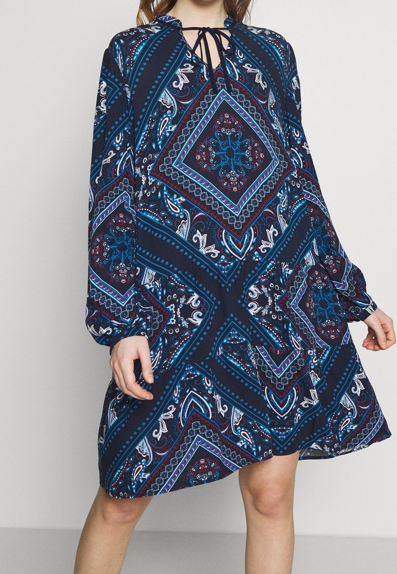 Långärmad klänning i marinblått med intrikata paisleymönster, som har geometriska former i rött och turkos, samt en nyckelhålshalsringning med snörning.