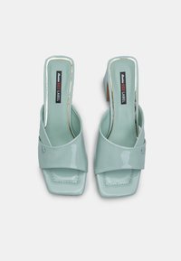 Light teal patent leather heeled sandals featuring a wide strap across the front, square toe, and a low block heel. Elegant and modern design.
