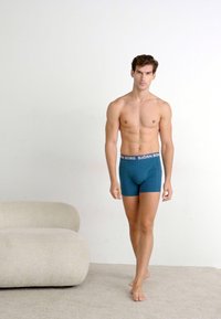 Boxer in teal con un elastico scuro con il nome del marchio "BJÖRN BORG." Il modello si trova su una superficie beige morbida davanti a uno sfondo chiaro.