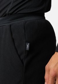 Zwarte fleecebroek met een gestructureerde afwerking en een ribbelband, voorzien van een klein merklabel aan de zijkant. Close-up weergave.