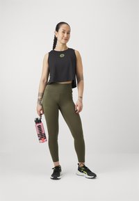 Svart avkortet tanktop med grønn aksent, grønne høytlivs leggings, rosa vannflaske, svarte joggesko med grønne detaljer, nøytral bakgrunn.
