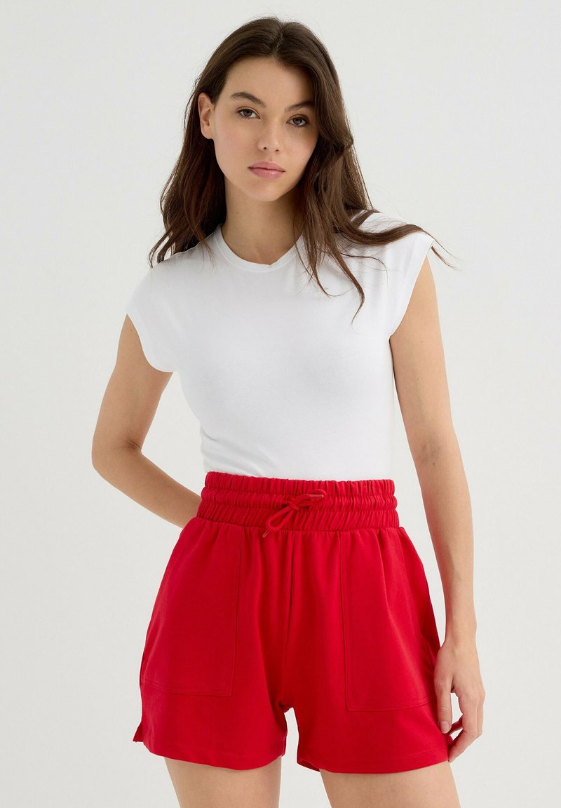 Pantalones cortos rojos de cintura elástica con bolsillos laterales, combinados con una camisa ajustada de manga corta blanca, sobre un fondo neutro.
