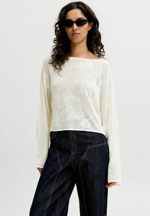 Mujer con cabello rizado que lleva gafas de sol negras, una blusa blanca de punto suelto de manga larga y vaqueros oscuros de talle alto y pierna ancha.