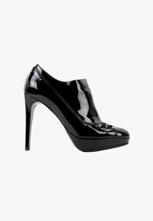 Chaussure à talons hauts noire brillante avec semelle compensée et boucle métallique décorative sur le dessus, vue de profil.