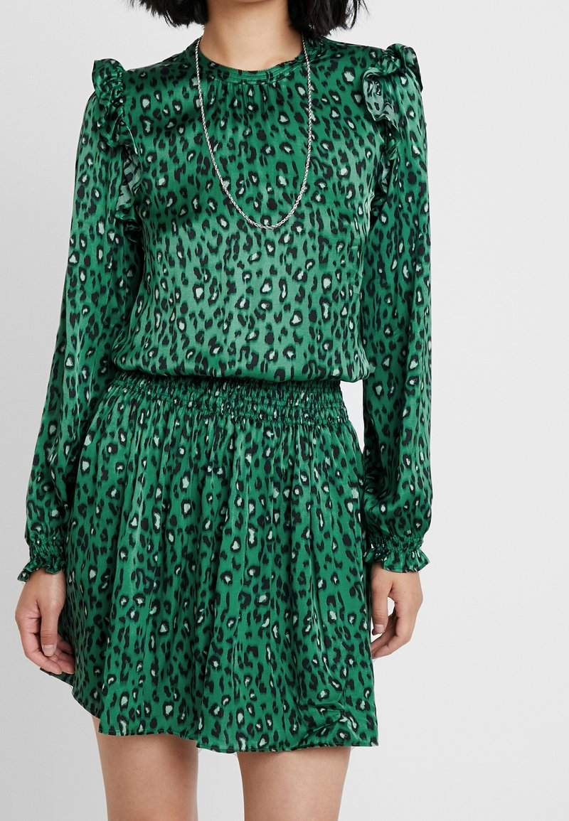 Robe verte à motifs léopard avec manches longues, accents d'épaules volantés, taille élastique et texture de tissu lisse. Détail d'un collier en argent visible.