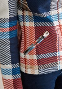Gros plan d’une veste à carreaux rouge, bleue, beige et blanche avec une poche zippée inclinée et une texture de tissu visible.