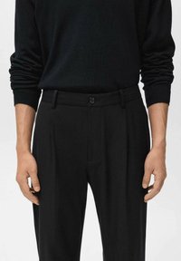 Persoon in een zwart shirt met lange mouwen, in de hoge zwarte broek met knoopsluiting en riemlussen gestoken.