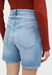 Ανοιχτό μπλε τζιν σορτς με πίσω τσέπες, φορεμένα από άτομο με μακρυμάνικη μπλούζα σε σκουριά χρώματος, εμφανίζεται από τη μέση και κάτω.