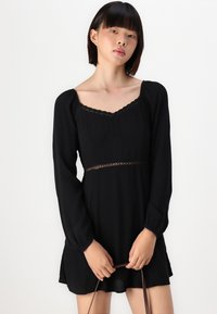 Melnas garām piedurknēm kleitas ar kvadrātveida dekoltē, teksturētu audumu, viļņotu apdari un saplacinātu vidukli, rotātu ar dekoratīvu mežģīni.