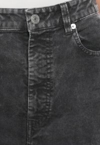 Jupe en velours côtelé noir avec une texture à nervures verticales, dotée d'un bouton à l'avant et de poches. Des détails en métal accentuent le design.