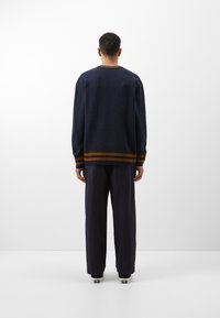 Marinblå tröja med rund halsringning, ribbade ärmslut och en kontrasterande randad nederkant i orange-brunt, kombinerad med lösa mörka byxor.