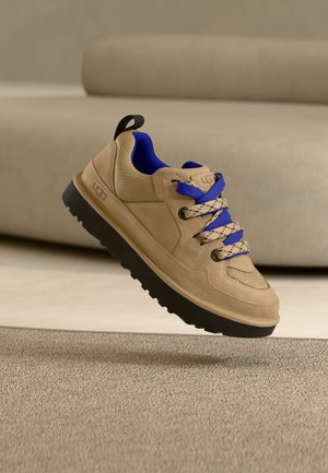 Baskets UGG beiges avec semelle noire et intérieur bleu, lacets bicolores bleus et beiges, flottant au-dessus d'un tapis beige texturé.