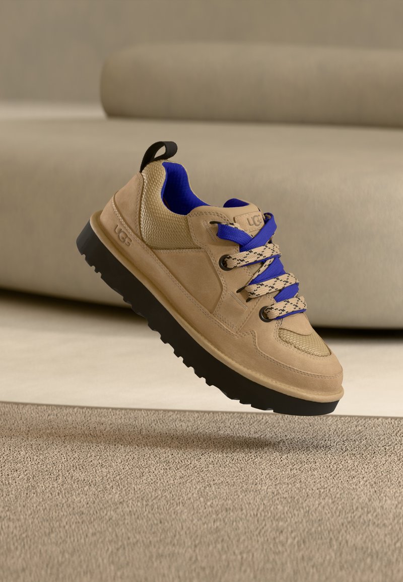 Baskets UGG beiges avec semelle noire et intérieur bleu, lacets bicolores bleus et beiges, flottant au-dessus d'un tapis beige texturé.