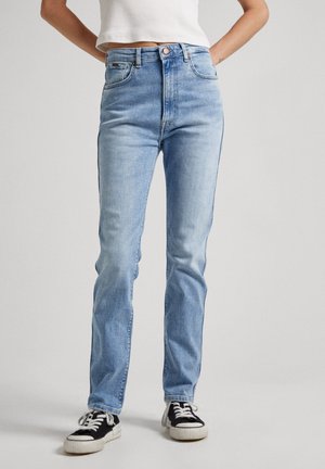 Hele sinised kõrge vöökohtadega teksad, mis on valmistatud denimist. Iseloomustab klassikaline viie tasku disain ja peen kulumine. Kombineeritud mustade ja valgete tossudega.