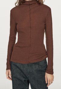 Brown long-sleeve turtleneck top made of a soft material, featuring a central seam detail and a fitted silhouette. Paired with dark denim jeans.
