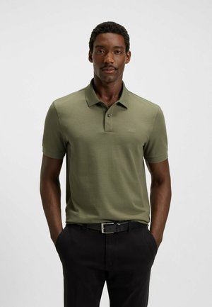 Olivenfarbener Poloshirt mit kurzen Ärmeln und feiner Textur, Kragen mit drei Knöpfen und Logodetail auf der Brust, kombiniert mit schwarzen Hosen und Gürtel.