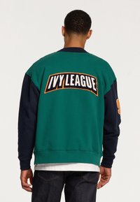Zelená a námořnická mikina s vyšitým logem "IVY LEAGUE" na zadní straně. Hladké manžety a lem, volný střih, měkká struktura.