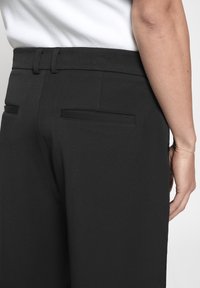 Pantalons noirs en tissu lisse, avec une taille haute, deux poches arrière et une coupe ajustée, vus de côté.