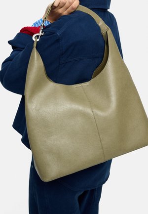 Oliwkowo-zielona skórzana torba na ramię z pojedynczym paskiem, noszona przez osobę w granatowej kurtce i dopasowanych spodniach.