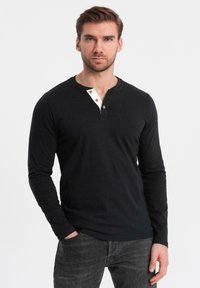 Schwarzes Langarmshirt mit weißer Knopfleiste, die aus drei braunen Knöpfen besteht. Glatte Textur mit Rundhalsausschnitt und taillierter Passform.