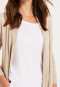 Ljusbeige, lätt stickad cardigan med öppen front, lång ärm och strukturerad yta, lagd över en enkel vit topp.