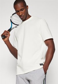 Vit bomullströja med kort ärm och rundad halsringning, ihop med grå svettbyxor. En tennisracket hålls över axeln.