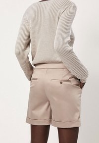 Person trägt einen beige gerippten Pullover und passende beige maßgeschneiderte Shorts mit Taschen, steht mit den Händen in den Taschen und blickt weg.