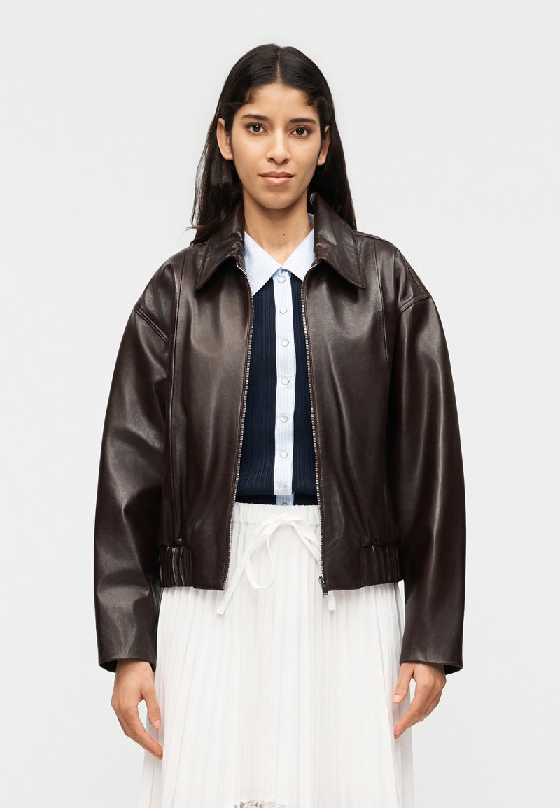 Γυναίκα που φοράει ένα σκούρο καφέ δερμάτινο μπουφάν πάνω από ένα μπλε ναυτικό και λευκό πουκάμισο με κουμπιά και μια λευκή πλισέ φούστα, στέκεται μπροστά σε έναν απλό φόντο.