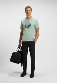Light green cotton t-shirt with a circular graphic design, paired with black tapered pants and carrying a black duffel bag.
