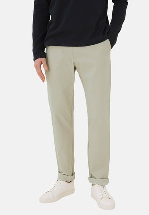 Homme portant un pantalon beige clair retroussé, des baskets blanches et une chemise noire à manches longues, les mains dans les poches, debout devant un fond blanc.