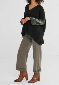 Femme portant un poncho noir à col en V avec des manches à motifs, un pantalon olive retroussé et des bottines marron à talons, debout les mains jointes.