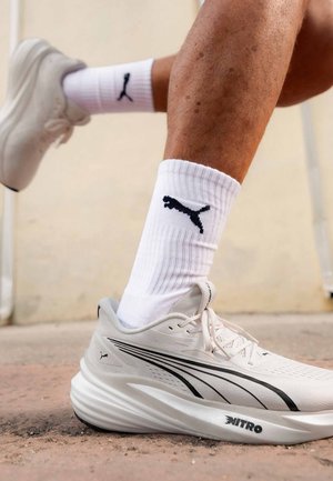 Osoba nosząca białe buty do biegania Puma Nitro oraz białe skarpetki do połowy łydki z czarnym logo Puma, stojąca na teksturowanej powierzchni na świeżym powietrzu.