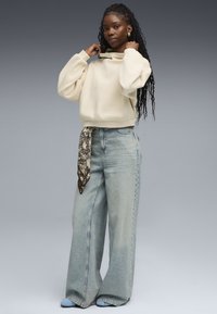Sudadera de color beige claro y corte oversize con un cuello alto, combinada con pantalones vaqueros de pierna ancha de color azul claro. Se lleva una bufanda estampada atada a la cintura.