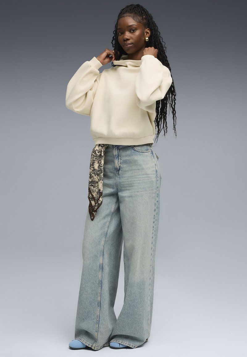 Sudadera de color beige claro y corte oversize con un cuello alto, combinada con pantalones vaqueros de pierna ancha de color azul claro. Se lleva una bufanda estampada atada a la cintura.