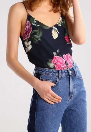 Vrouw die een donkerbloesem top zonder mouwen draagt, ingestopt in een high-waisted blauwe spijkerbroek, met één hand in een zak rustend.