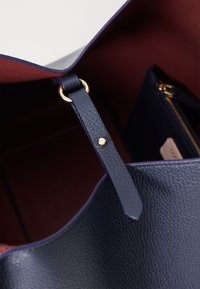 Navy leather tote bag with a textured finish, featuring a gold-tone hardware detail on a strap and a contrasting burgundy interior.