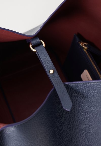 Sac fourre-tout en cuir marine avec une finition texturée, présentant un détail en matériel doré sur une sangle et un intérieur bordeaux contrastant.