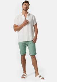 Weißes Kurzarmhemd aus strukturierter Baumwolle, kombiniert mit hellgrünen hochgekrempelten Shorts und weißen Sandalen. Schlichtes Design, lässige Passform.