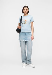 Femme portant un t-shirt à imprimé floral bleu, une jupe en jean par-dessus un pantalon en jean, des baskets blanches et un sac à bandoulière noir.