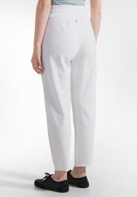 Kvinde iført højtaljede hvide bukser og sorte balletsko, stående mod en ensfarvet hvid baggrund, vendt væk fra kameraet.