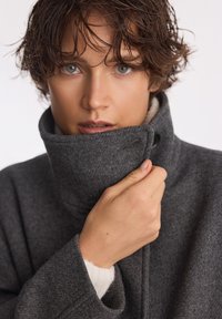 Szary wełniany płaszcz z wysokim kołnierzem, posiadający miękką podszewkę i boczne kieszenie. Model trzyma kołnierz zapięty jedną ręką.