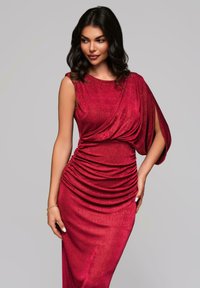 Abito rosso aderente con tessuto texturizzato, spalla drappeggiata asimmetrica e dettagli arricciati in vita, creando una silhouette lusinghiera.