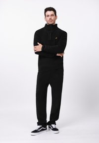 Juodas vilnonis megztinis su aukštu apykakle ir užtrauktuku, derinamas su atitinkančiomis juodomis kelnėmis. Ant krūtinės ryškus geltonas logotipas. Tekstūruota medžiaga.
