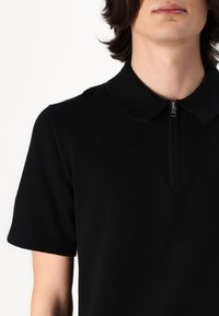 Svart polo-skjorte med korte ermer, flat krage og en kvart lengde glidelås. Stoffet har en glatt, teksturert overflate.