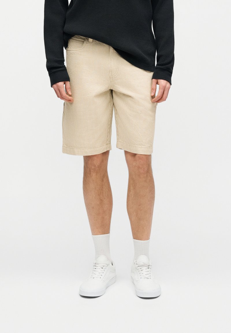 Man draagt beige korte broek met krijtstreepjes, zwart shirt met lange mouwen, witte sokken en witte sneakers, staand tegen een effen achtergrond.