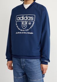 Mörkblå Adidas tröja med V-ringning, med en stor vit logotyp och textdesign, samt elastiska manschetter. Bärs över denimbyxor.