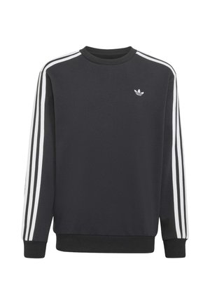 Ung dreng iført en sort Adidas sweatshirt med hvide striber på ærmerne og blå Adidas jeans med striber langs siderne.