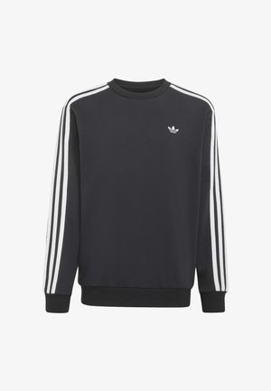 Młody chłopiec w czarnej bluzie Adidas z białymi paskami na rękawach i niebieskich dżinsach Adidas z paskami po bokach.