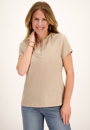 Lachende vrouw met lang haar, die een beige shirt met korte mouwen draagt, blauwe spijkerbroek, gouden oorbellen in creoolse stijl en armbanden tegen een effen achtergrond.
