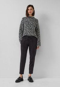 Kobieta ubrana w szaro-czarny sweter w leoparda, czarne zwężane spodnie i czarne loafersy, stojąca na tle gładkiej, jasnoszarej ściany.